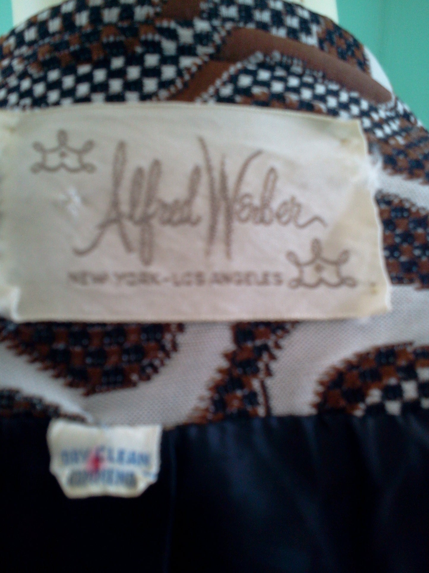 Close-up of a vintage patterned coat garment label reading Alfred Weber and New York and Los Angeles, with a small 'GUESS CLEAN GUESS' tag below it. The fabric has a black, white, and brown geometric pattern.