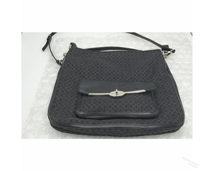 A black Coach jacquard crossbody handbag with a subtle geometric monogram pattern, featuring a front flap pocket secured by a silver-tone metal bar clasp, resting on a white textured surface.