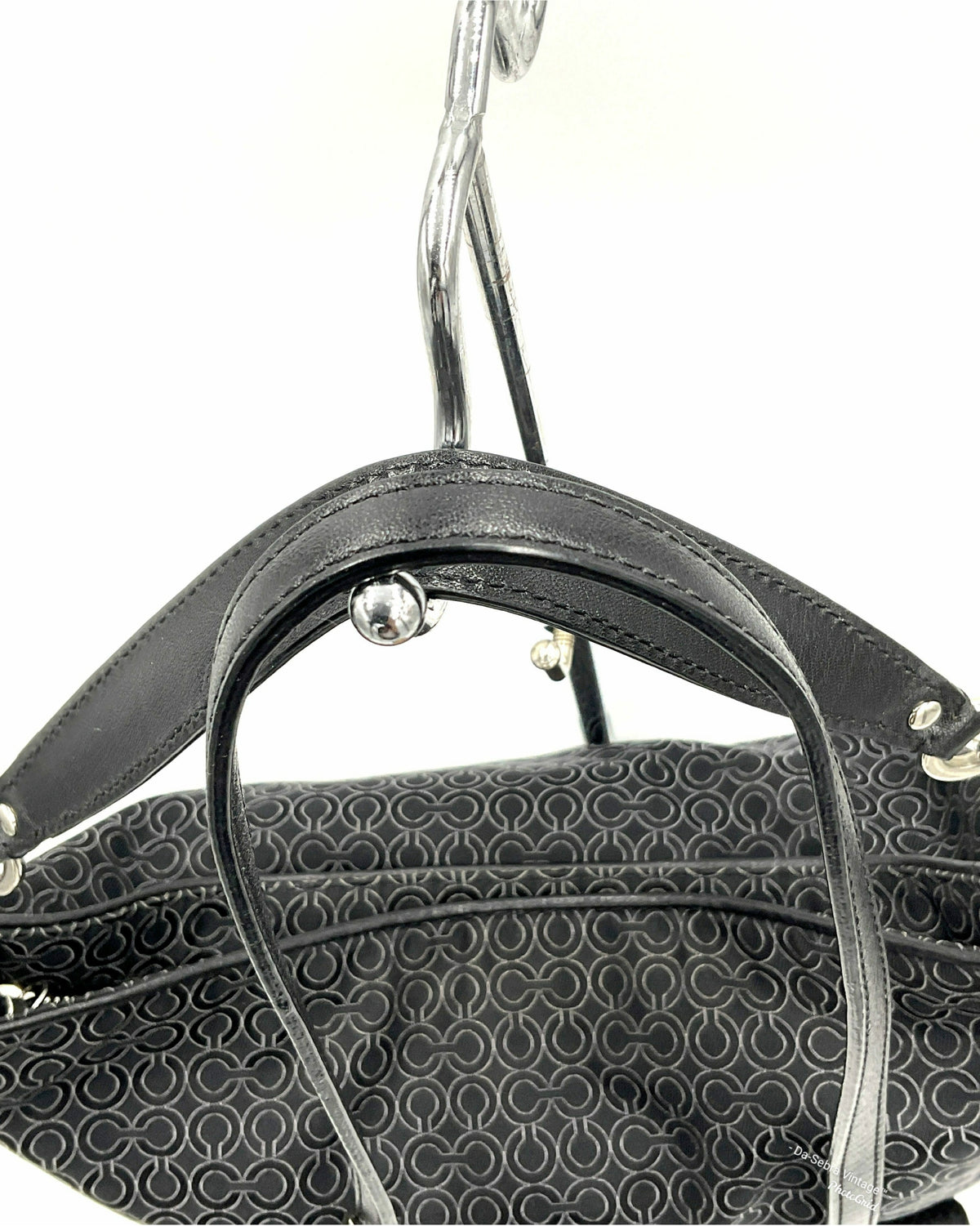 Close-up of a Coach black jacquard handbag handle with silver-tone hardware, hanging against a plain white background. The bag body features a dark gray and black repeating monogram pattern.