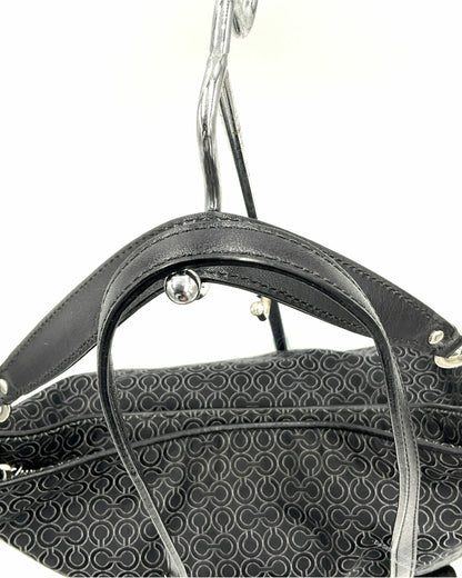 Close-up of a Coach black jacquard handbag handle with silver-tone hardware, hanging against a plain white background. The bag body features a dark gray and black repeating monogram pattern.