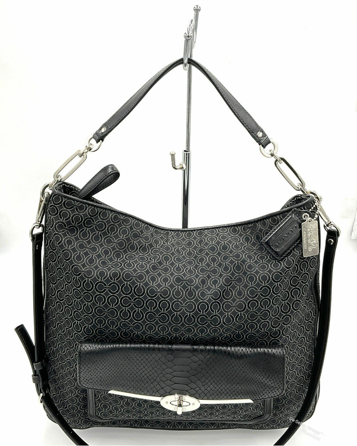 A Coach black jacquard hobo shoulder bag with a tonal monogram pattern, a front flap pocket with a silver-tone turn-lock closure, and silver hardware, shown hanging on a display stand against a white background.
