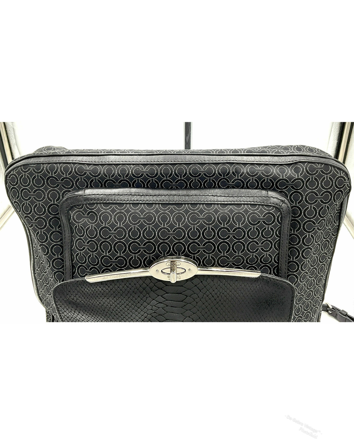 A close-up of a black jacquard Coach handbag featuring a textured geometric pattern on the main body and a front zip pocket with a silver-tone turn-lock clasp, set against a white background.