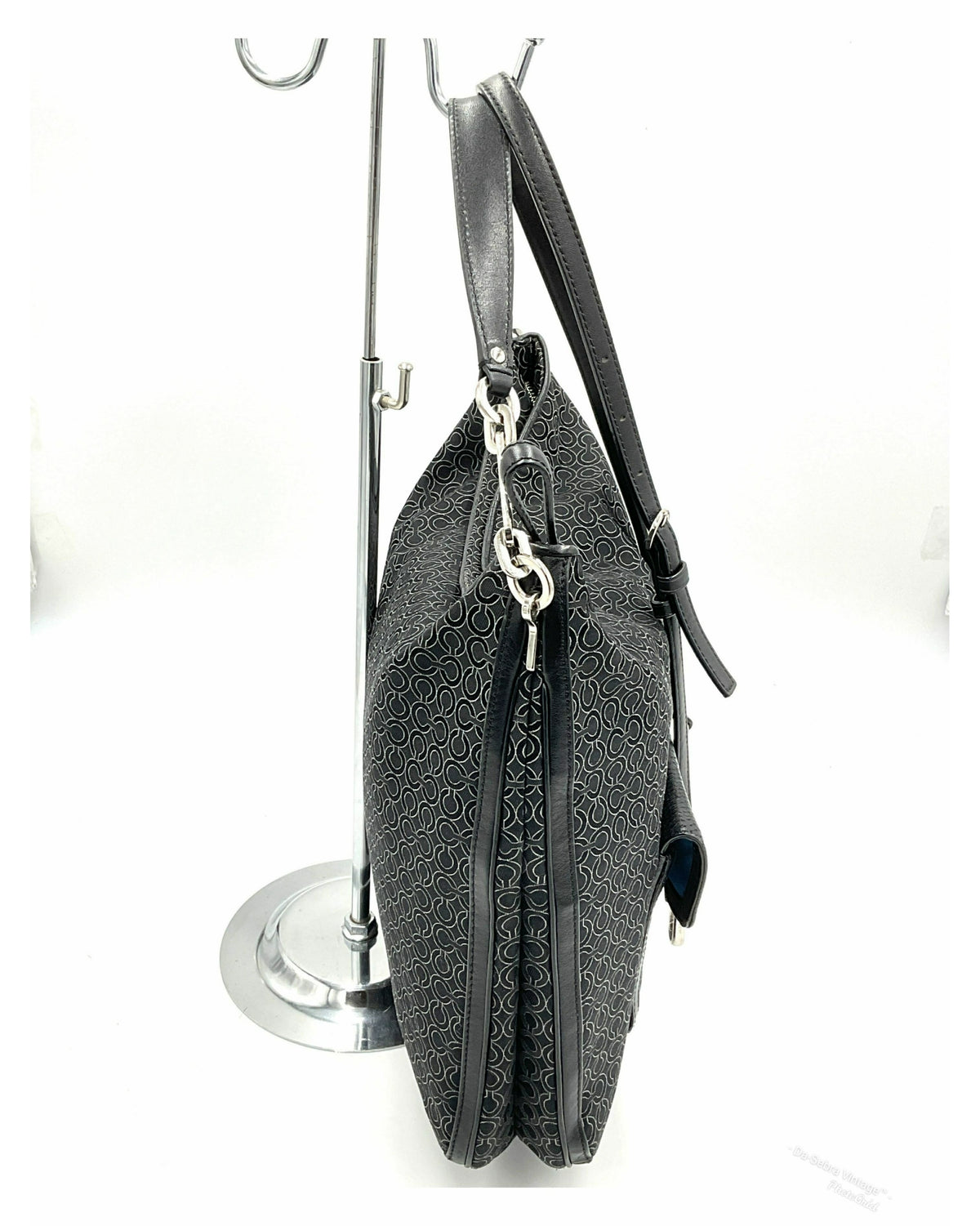 Side view- A Coach black jacquard crossbody handbag with a repeating monogram pattern, leather trim, displayed on a silver metal stand against a white background.