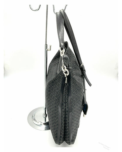 Side view- A Coach black jacquard crossbody handbag with a repeating monogram pattern, leather trim, displayed on a silver metal stand against a white background.