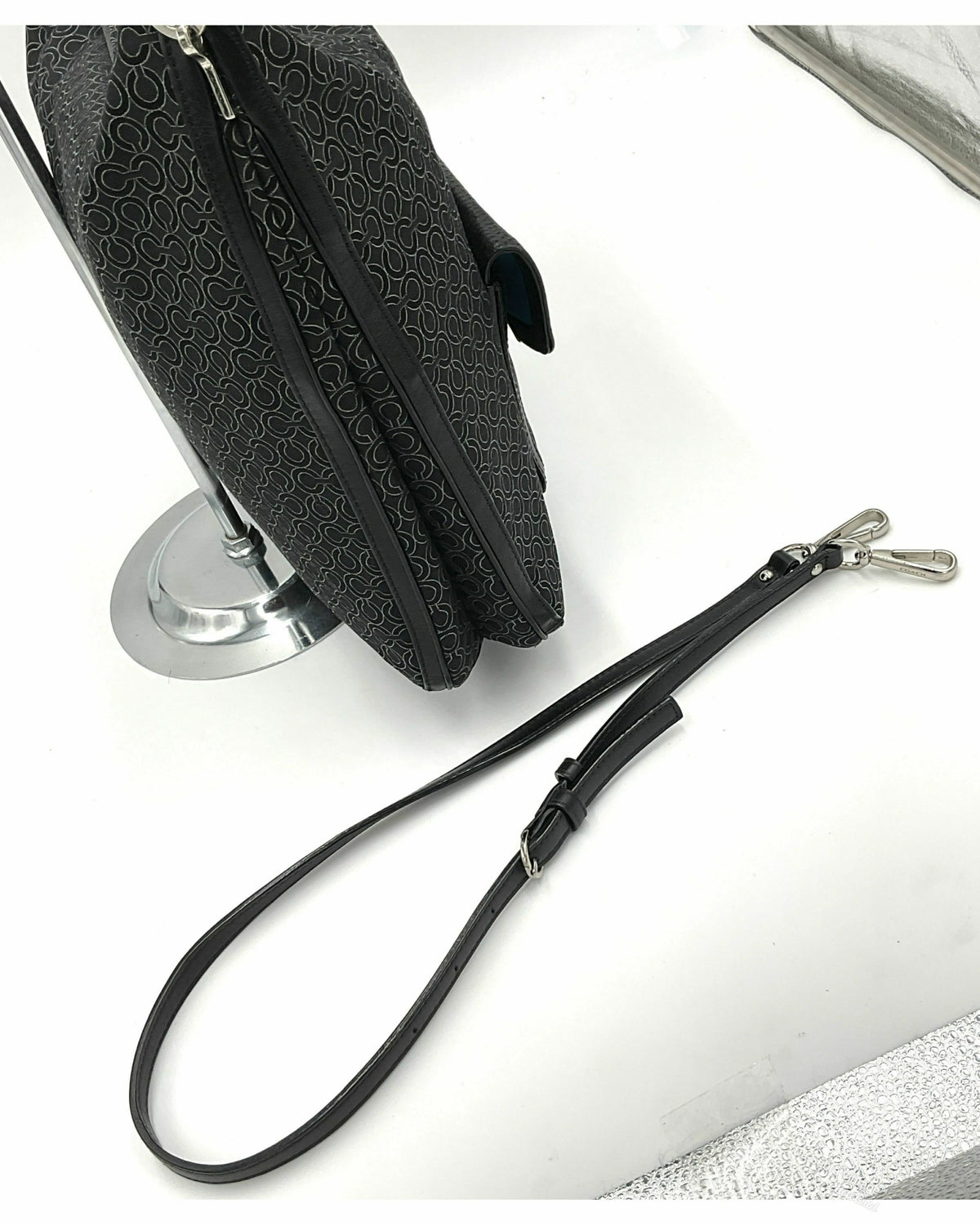 A Coach black jacquard patterned handbag with a detachable black leather wristlet strap and silver-tone hardware, displayed on a white surface next to a silver stand.
