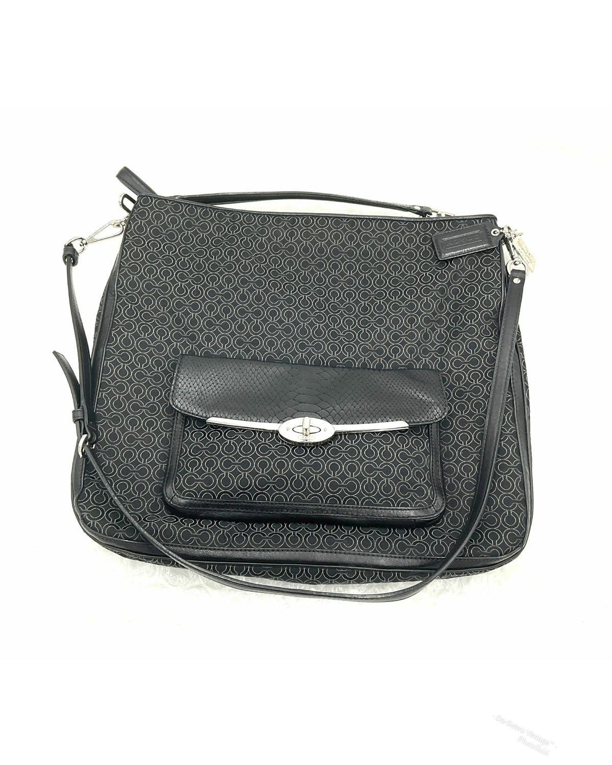 A black jacquard Coach crossbody handbag with a repeating monogram pattern, a front flap pocket with a silver-tone turn-lock closure, and a top handle, shown on a white background.