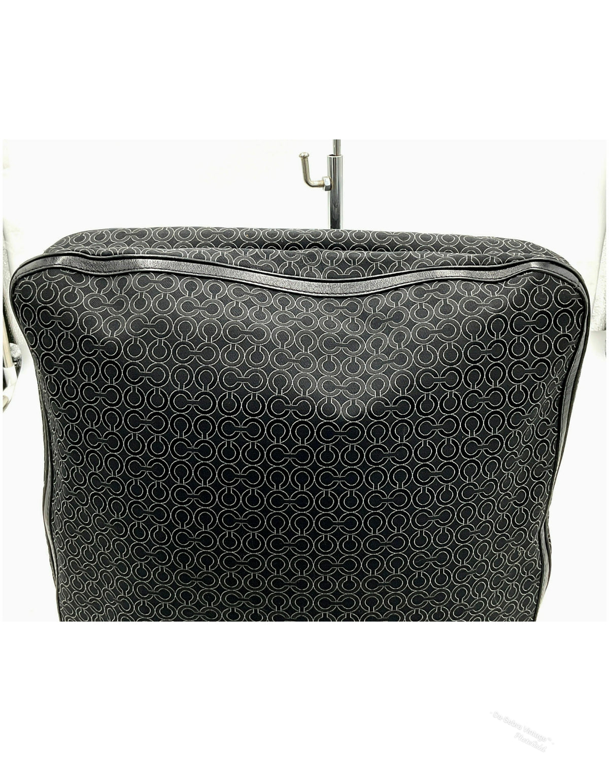 A black jacquard Coach bag with a repeating white monogram pattern, featuring a top zipper and a metal hanger hook inserted at the top, set against a plain white background.