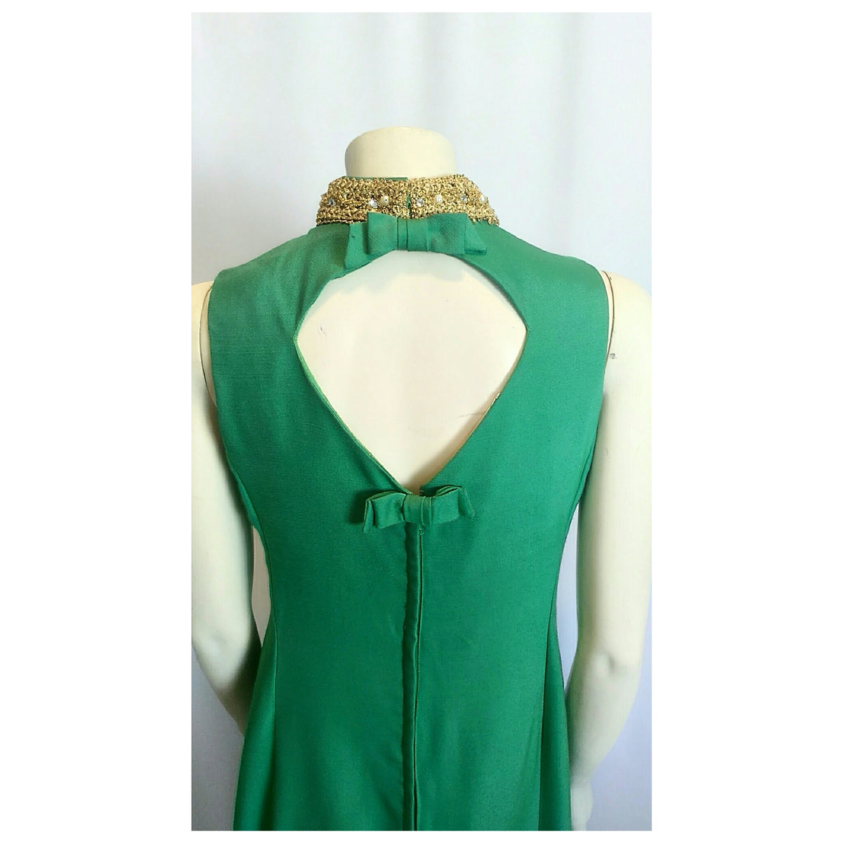 A green sleeveless dress displayed on a cream mannequin, showing the back view with a large keyhole cutout, bow detail, and a gold beaded collar.