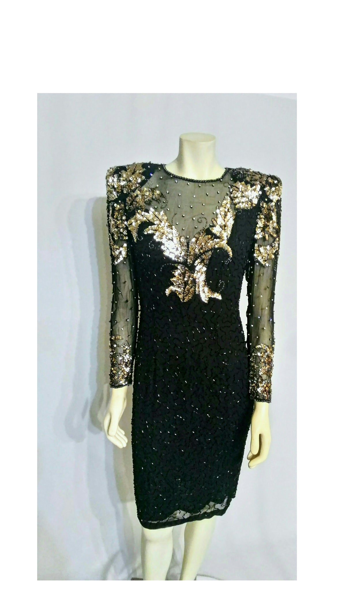 A black sheer embellished long-sleeve  dress with gold, silver sequins and rhinestones.   Hand-sewn black and clear bugle beading, and glittering crystal rhinestones,  displayed on a mannequin against a light gray background.