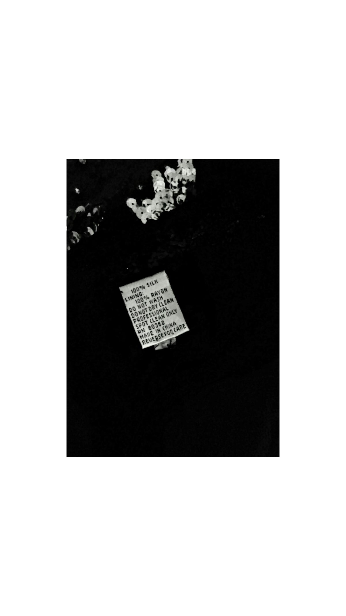 The inside of black dress, featuring a small white printed label with text and a cluster of small white sequins near the upper left chest area, shown against a plain white background.