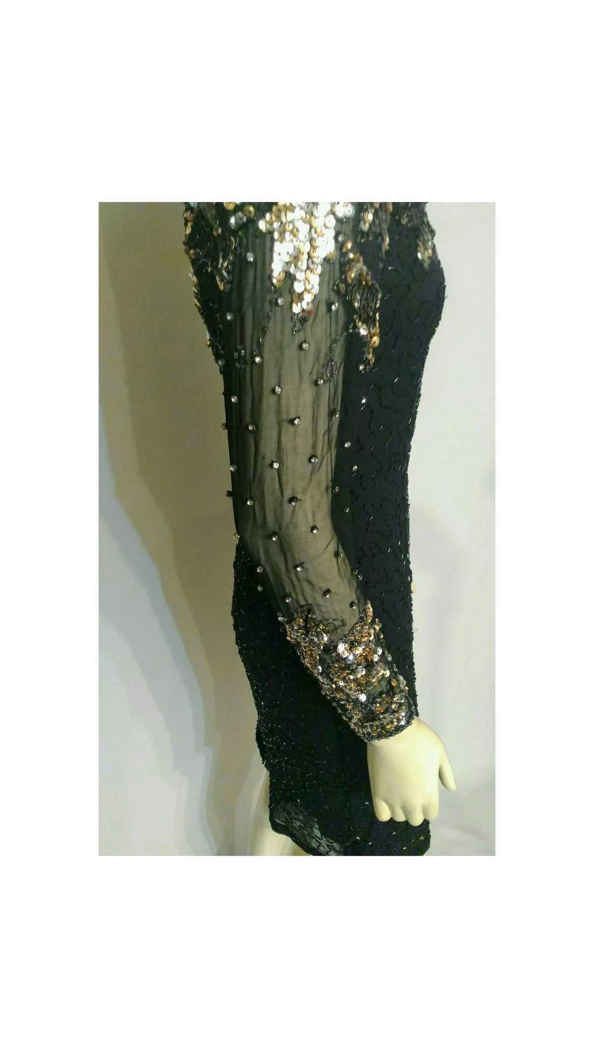 A close-up of a black sequined dress or garment displayed on a mannequin, featuring sheer mesh sleeves with gold sequin embellishments and rhinestones
