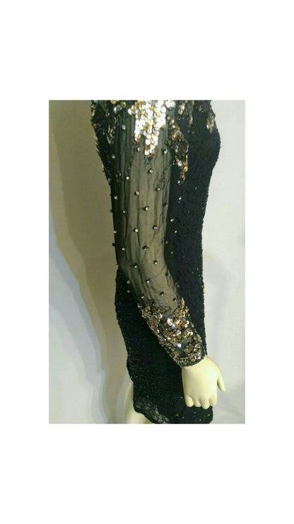 A close-up of a black sequined dress or garment displayed on a mannequin, featuring sheer mesh sleeves with gold sequin embellishments and rhinestones