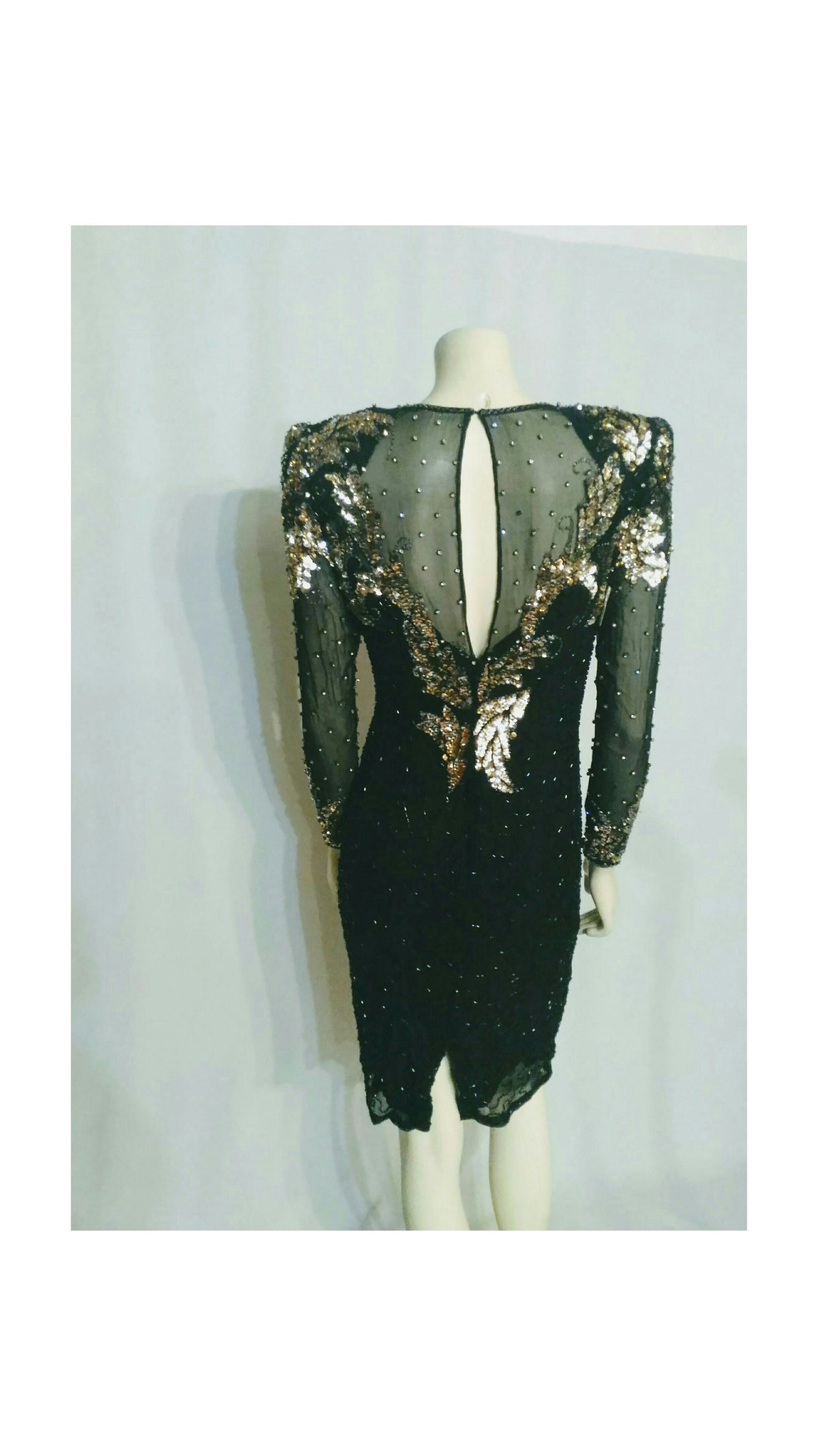 A black sequined evening dress with sheer mesh sleeves and a deep keyhole back, displayed on a mannequin against a light gray background.