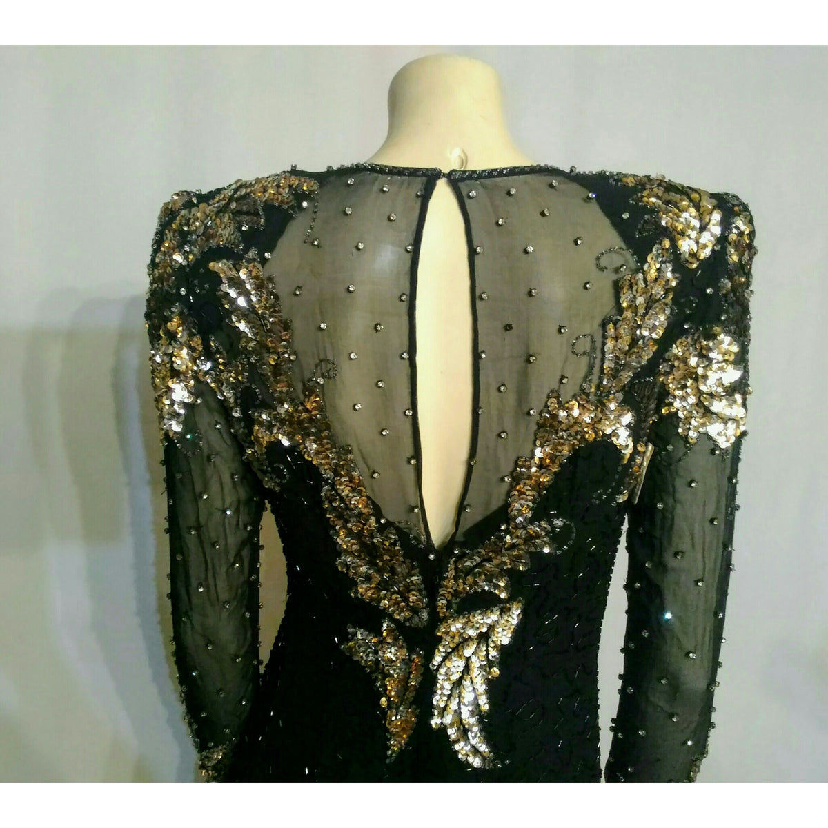 A black long-sleeve evening gown with a sheer back panel, embellished with gold sequins, beads, and rhinestones, displayed on a mannequin. No rhinestones are missing on the back and no snags or runs on sheer material