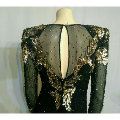 A black long-sleeve evening gown with a sheer back panel, embellished with gold sequins, beads, and rhinestones, displayed on a mannequin. No rhinestones are missing on the back and no snags or runs on sheer material