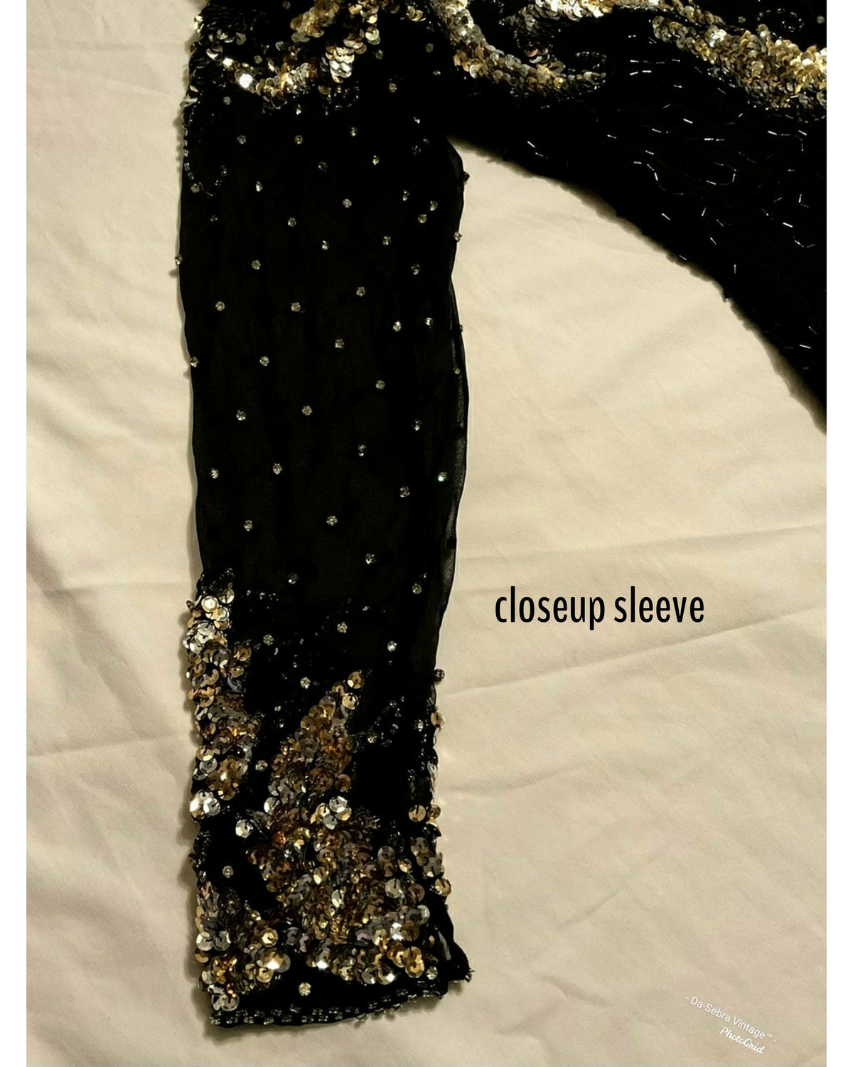 Close-up of a black embellished sleeve with rhinestones and a gold sequined cuff, laid flat on a white fabric background with the text 'closeup sleeve'. 