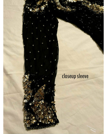Close-up of a black embellished sleeve with rhinestones and a gold sequined cuff, laid flat on a white fabric background with the text 'closeup sleeve'. 