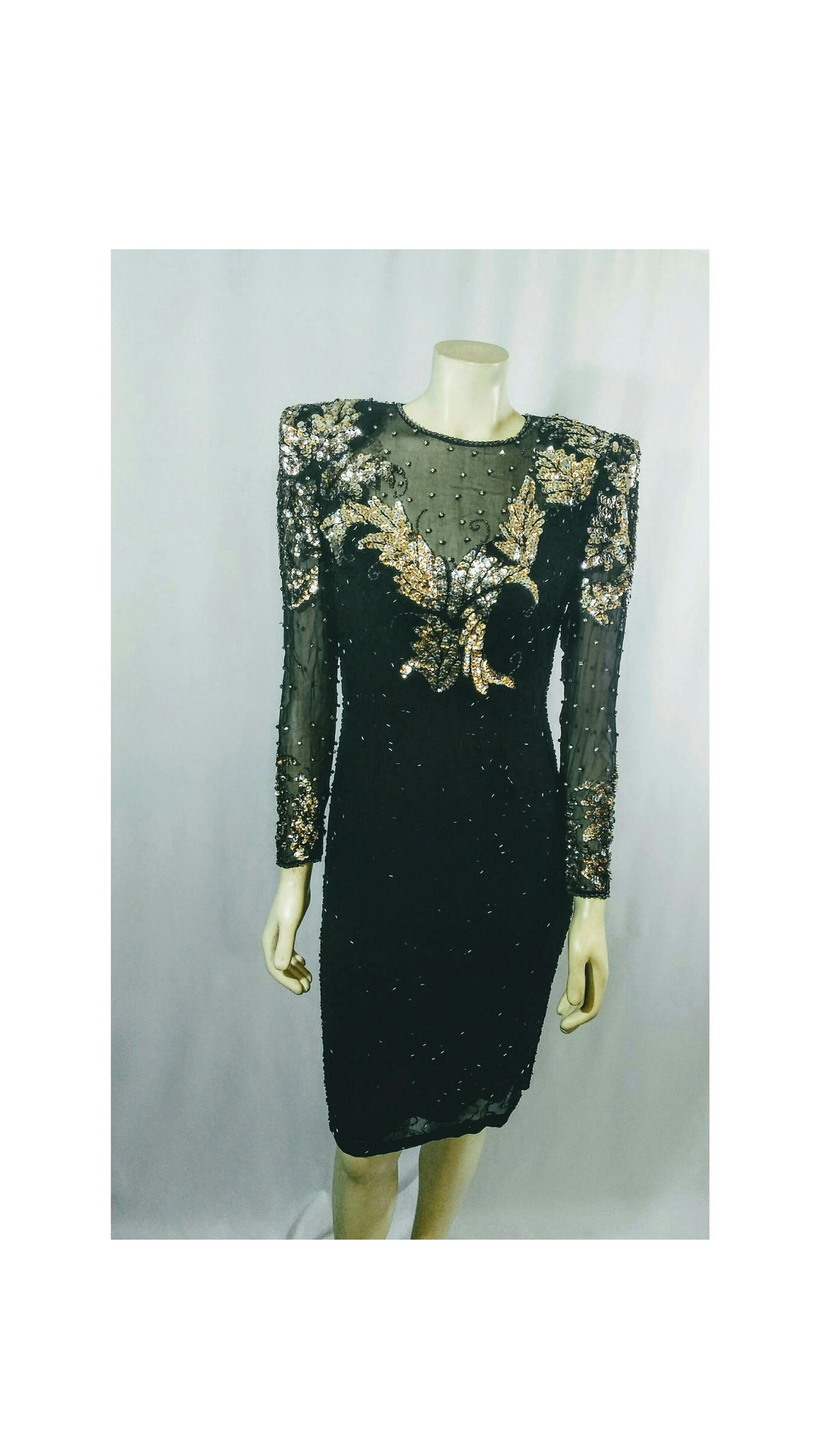 A black long-sleeve knee length dress with gold and silver sequin. Embellishments of rhinestones on the sheer bodice and sleeves, displayed on a mannequin against a light gray backdrop.