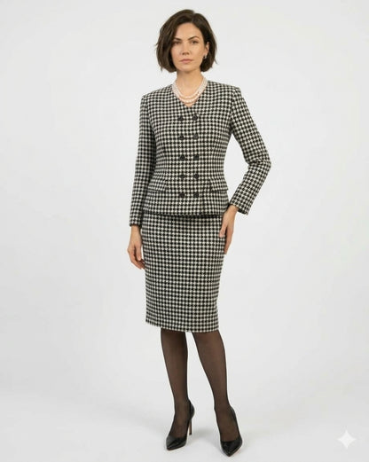 A woman modeling a black and white houndstooth suit set consisting of a double-breasted blazer with a pearl collar and matching pencil skirt, styled with sheer black tights and black heels against a light gray studio background.