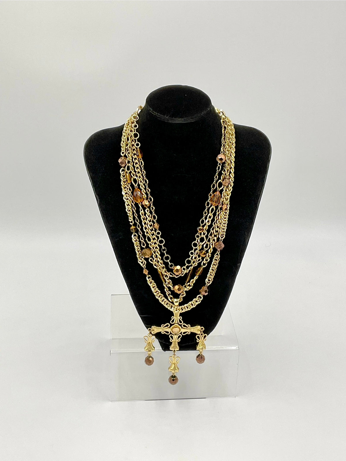 A multi-strand gold chain necklace with brown beads and a gold cross pendant, displayed on a black velvet jewelry bust stand.