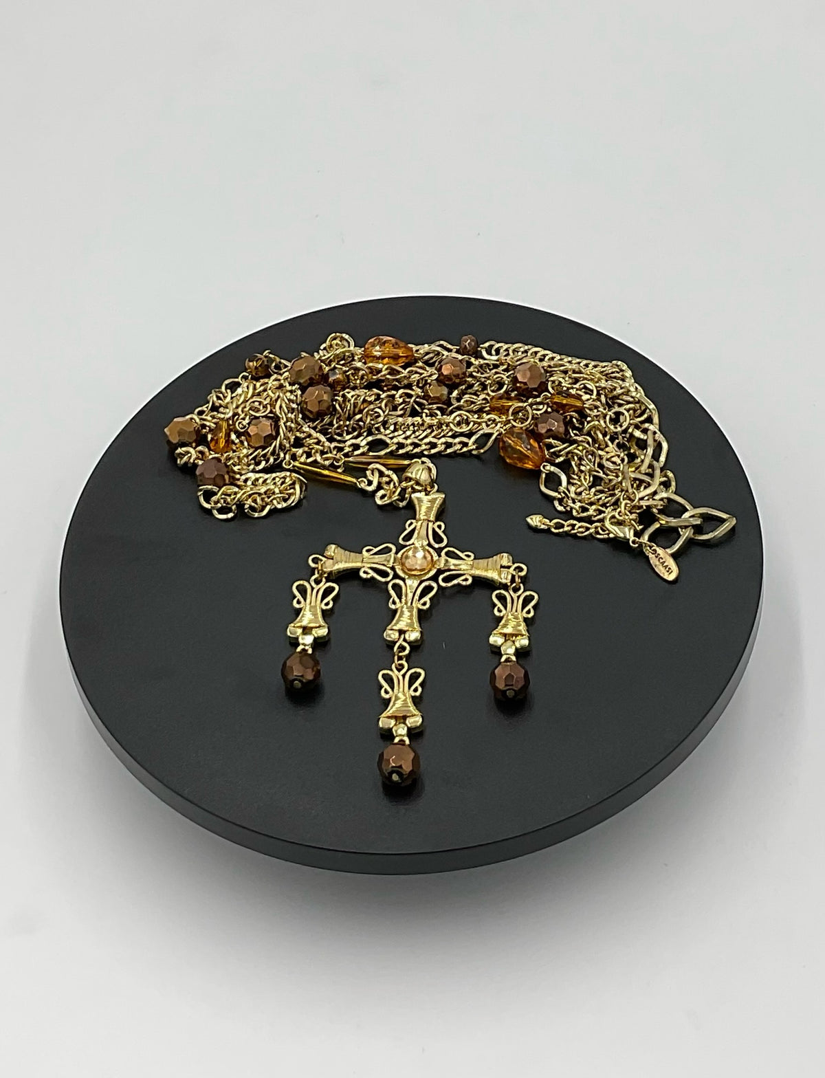 A gold-toned religious necklace and matching earrings with amber and brown beads, displayed on a round black stand against a light gray background.