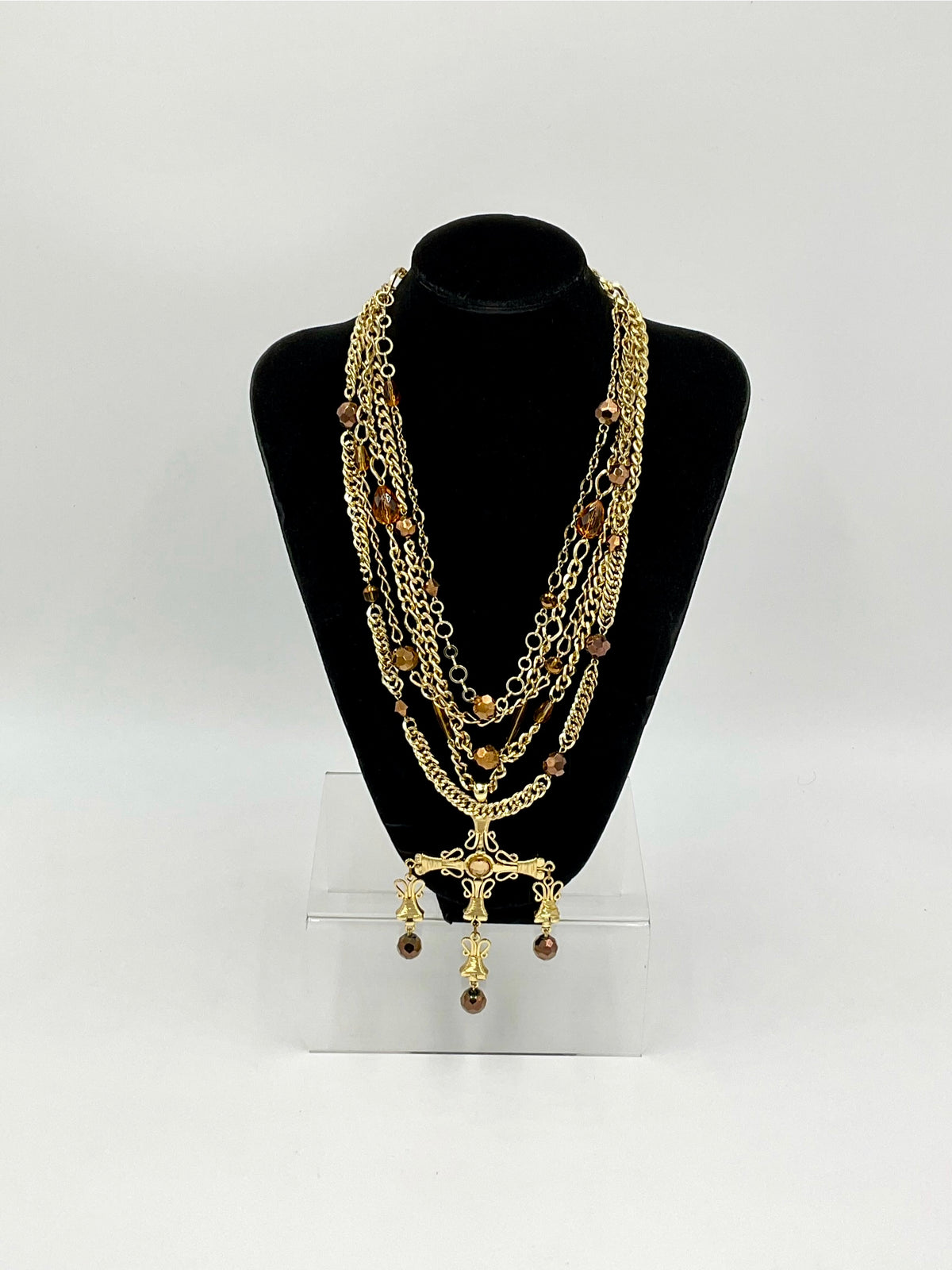 A multi-layered gold chain necklace with brown beads and a cross pendant, displayed on a black neck mannequin bust over a clear acrylic stand against a white background.