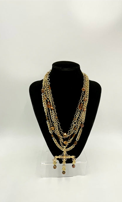 A multi-layered gold chain necklace with brown beads and a dangling cross pendant, displayed on a black neck mannequin bust against a white background.