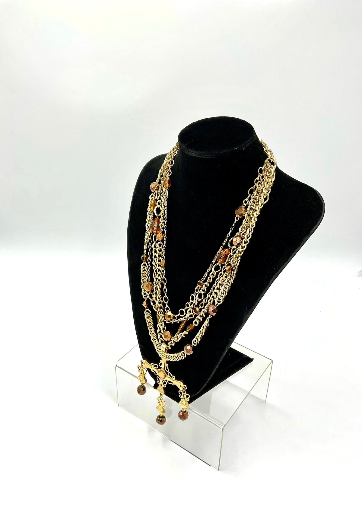 A multi-strand gold chain necklace with brown beads and charms, displayed on a black velvet jewelry bust stand on a clear acrylic riser against a white studio background.