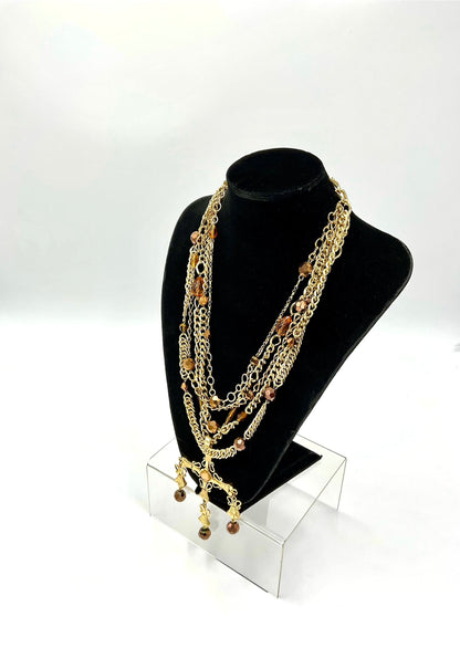 A multi-strand gold chain necklace with brown beads and charms, displayed on a black velvet jewelry bust stand on a clear acrylic riser against a white studio background.