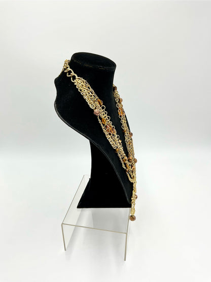 A gold-toned multi-strand chain necklace with amber-colored beads, displayed on a black velvet jewelry bust stand on a clear acrylic riser against a white studio background.