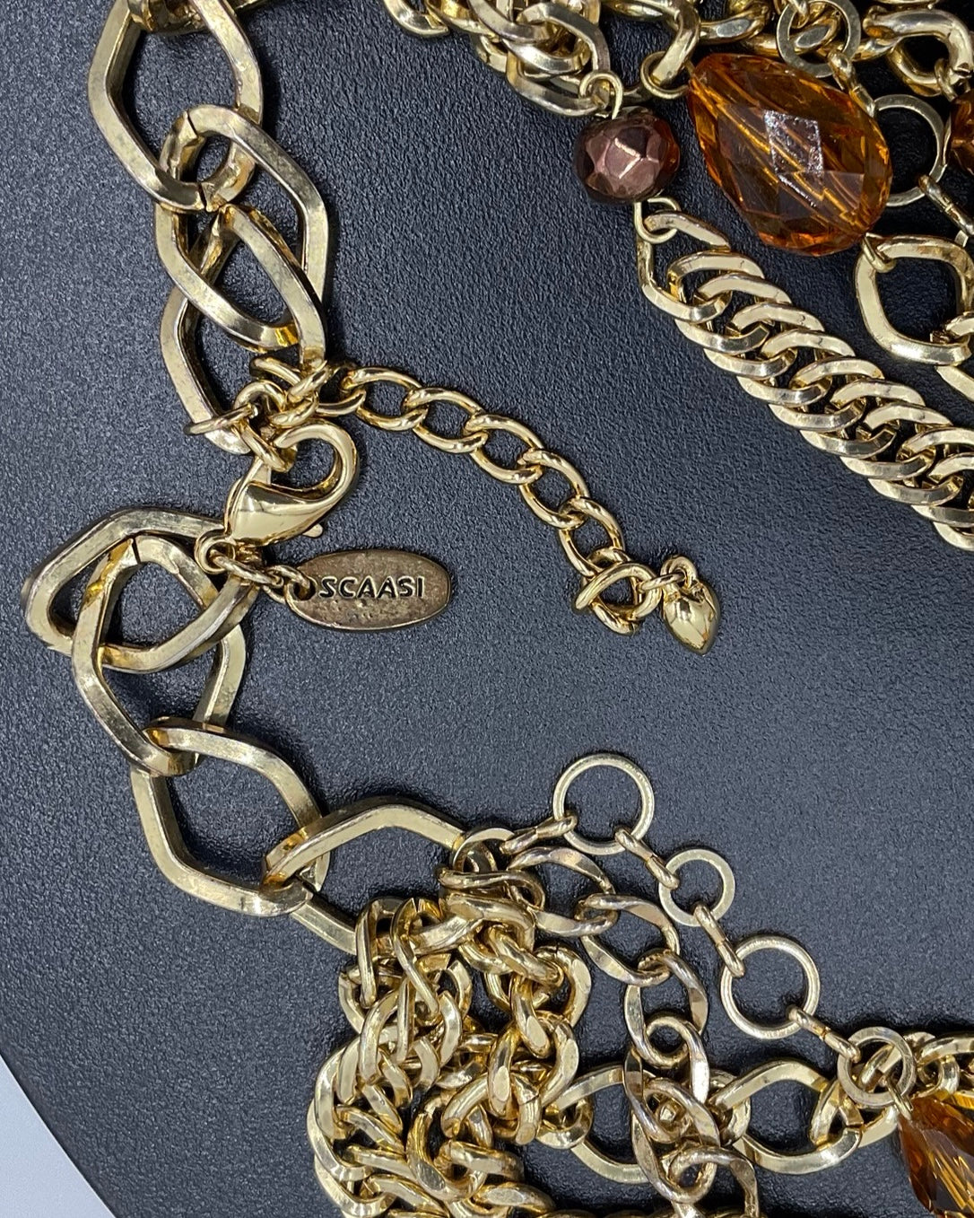 Close-up of a gold-toned chain necklace with chunky links, a lobster clasp, extender chain, and faceted amber-brown crystal beads on a dark textured background.