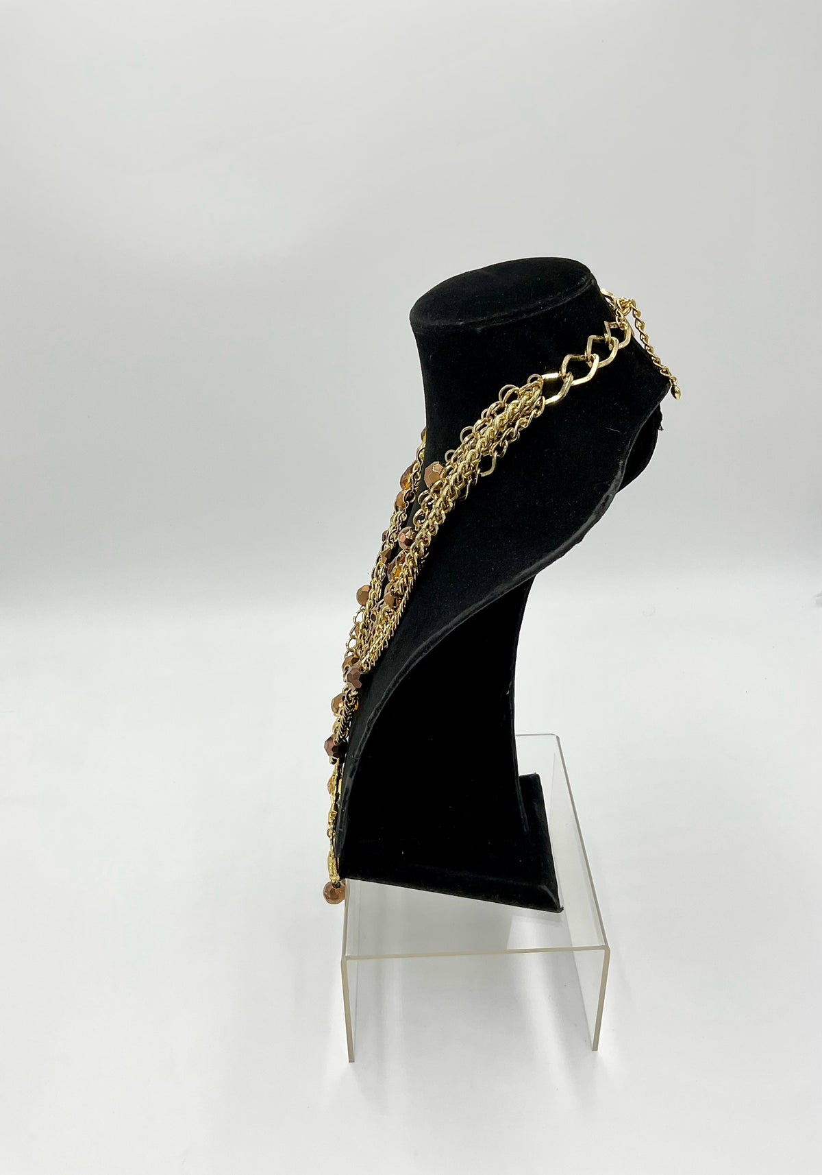 A gold-toned multi-strand chain necklace with small bead details, displayed on a black velvet jewelry bust stand on a clear acrylic riser against a white studio background.