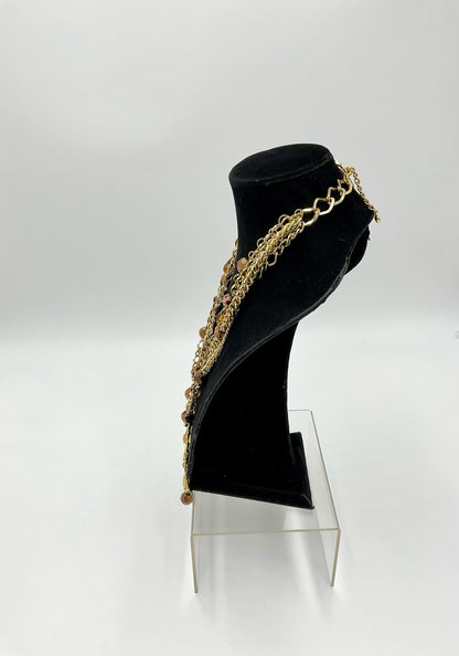 A gold-toned multi-strand chain necklace with small bead details, displayed on a black velvet jewelry bust stand on a clear acrylic riser against a white studio background.