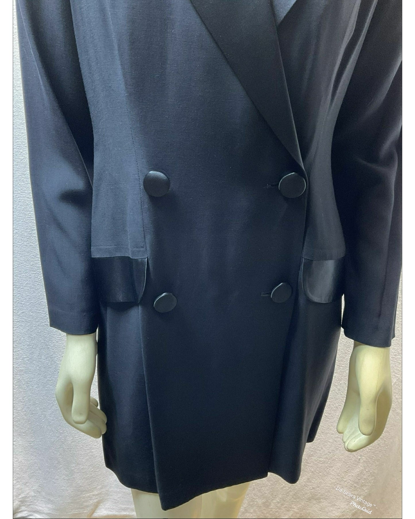 A black double-breasted Tuxedo style with wide lapels and large buttons, displayed on a mannequin with cream-colored hands against a light gray fabric backdrop.
