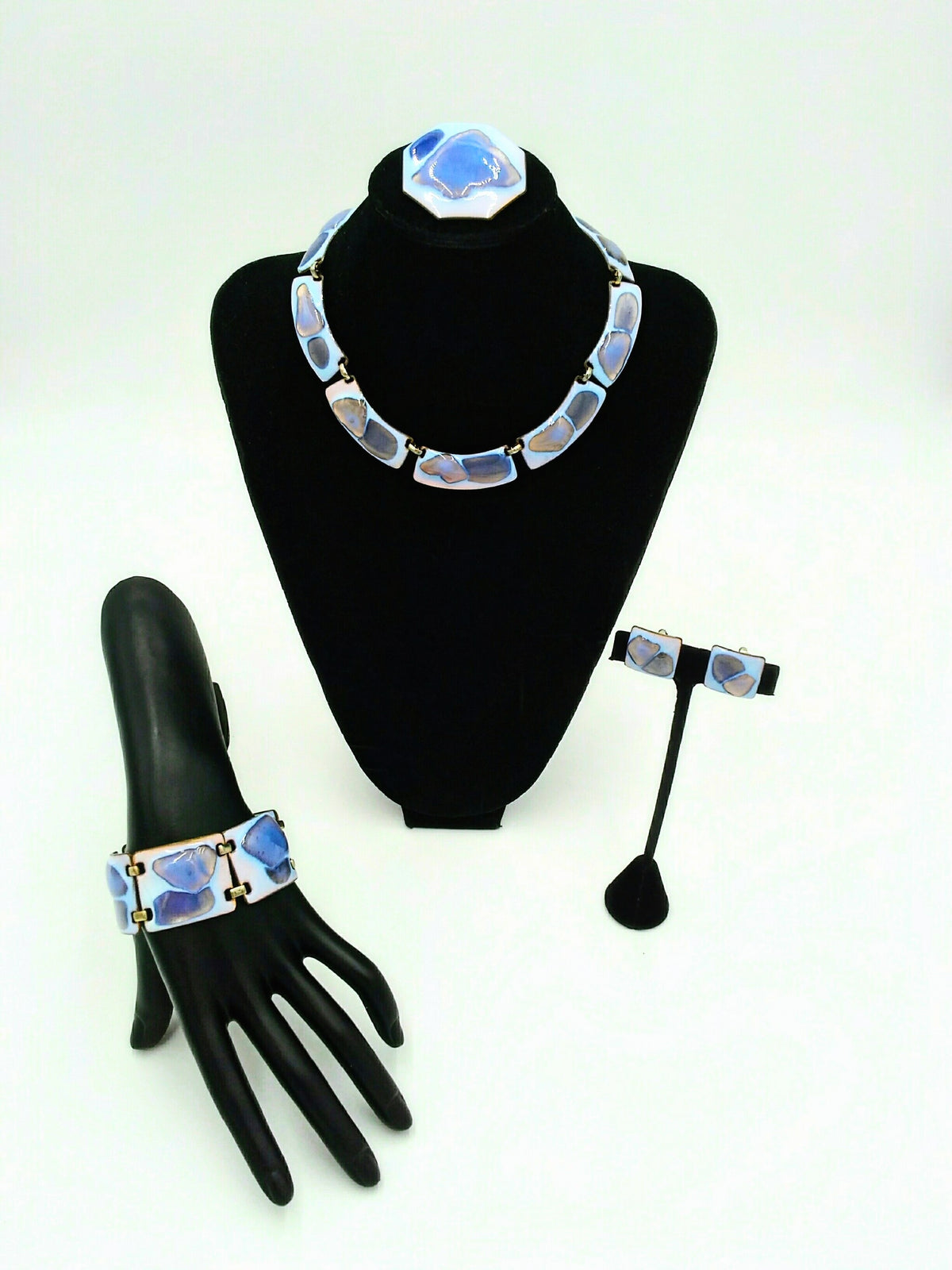 A jewelry set featuring a necklace, matching bracelet, earrings and brooch The color palette is exceptional: a striking opalescent blue enamel with beautiful hints of lavender and purple tones.  displayed on black mannequin stands against a white background.