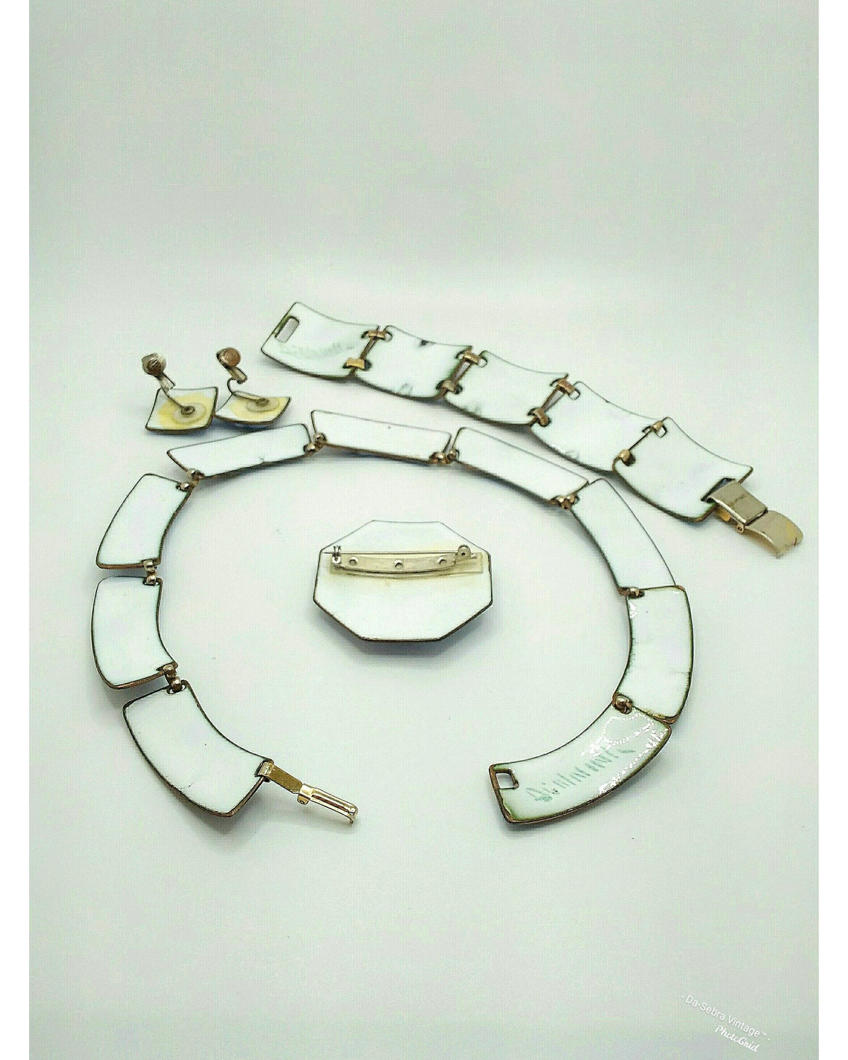 A vintage jewelry set featuring white enamel  geometric links with gold-tone metal settings, including a bracelet, necklace, earrings, and a pin, displayed on a light gray background.
